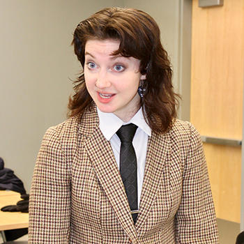 A woman with brown hair in a necktie and checked suit jacket talks to someone off camera.