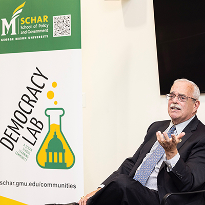 A man in a dark suit with gray hair and a moustache sits in front of a Democracy Lab banner.