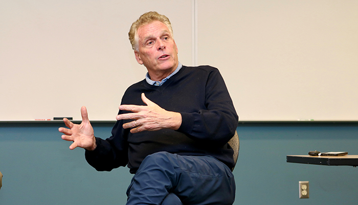 A man in a dark sweater against a whiteboard in the background gestures with his hands.