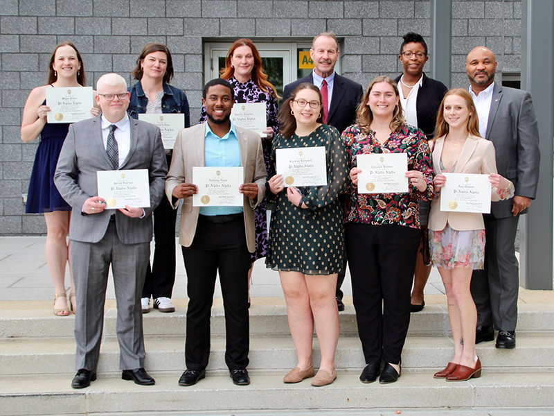 Ten young adults and a man in a gray suit hold certificates in front of them on a concrete staircase.
