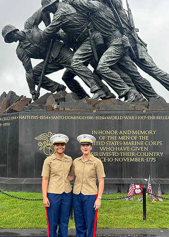 Two women in white military hats, tan shirts, and red and blue slacks pose in front of a statue.