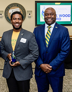 A man in a gray jacket holds a blue trophy while a man in a blue suit stands next to him.