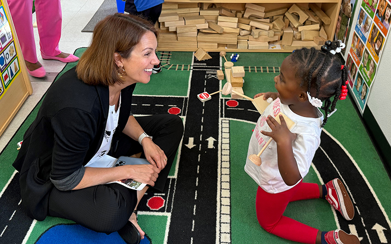 A woman in a dark sweater and white top sits on a green rug with a little girl in a white shirt.