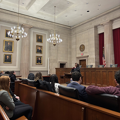 A group of students seated in rows listen to a woman wearing a black robe standing at a podium.