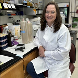 A woman in a white lab coat sits at a desk and smiles