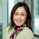 A woman in a gray shirt and scarf stands and smiles