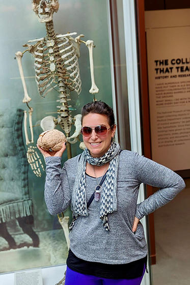 A smiling woman in a gray sweater and scarf standing in front of a skeleton holds a plasticized brain.