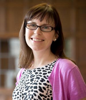 A woman in a black and white spotted top and wearing glasses smiles at the camera.